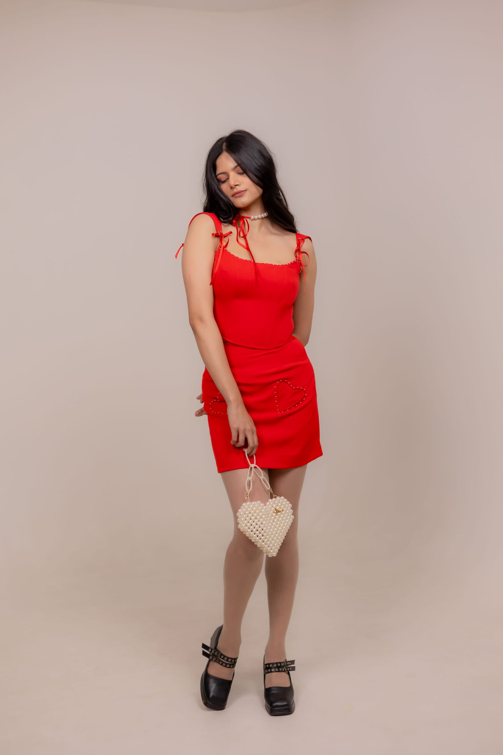 Full-length fashion portrait captured at Lens Grove Studio, a professional photo and video shoot rental space in Delhi, featuring a female model in a red dress holding a beaded handbag, styled against a minimal neutral backdrop with soft studio lighting for editorial and portfolio photography.