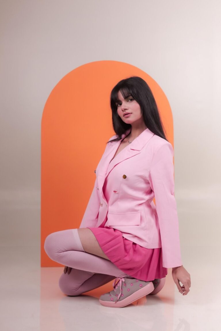 Editorial fashion portrait shot at Lens Grove Studio, a professional photo and video shoot rental space in Delhi, featuring a female model in a pink blazer and skirt seated against a bold orange arch backdrop, styled with clean studio lighting for fashion, portfolio, and brand photography.