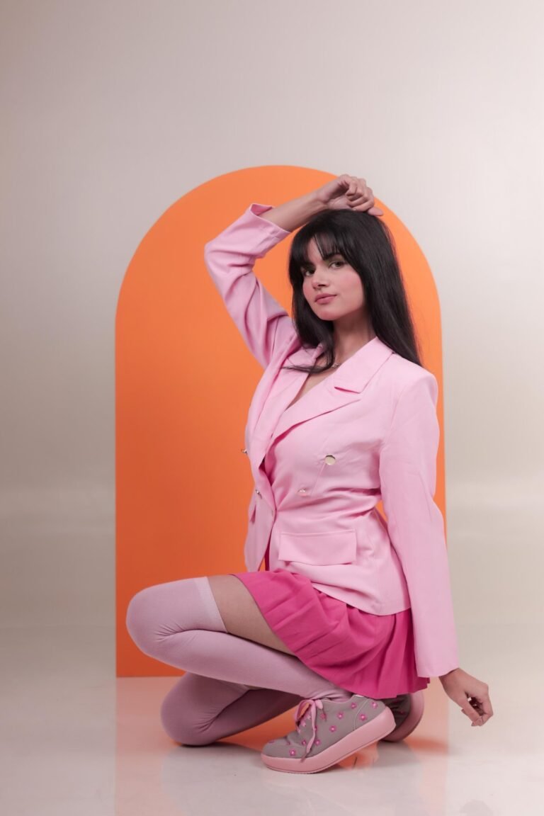 Editorial fashion portrait shot at Lens Grove Studio, a professional photo and video shoot rental space in Delhi, featuring a female model in a pink blazer and skirt seated against a bold orange arch backdrop, styled with clean studio lighting for fashion, portfolio, and brand photography.
