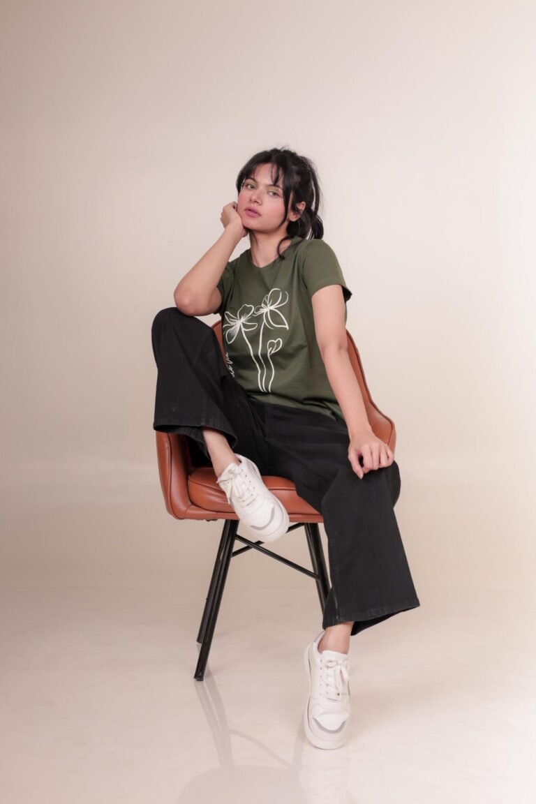 Casual studio portrait captured at Lens Grove Studio, a professional photo and video shoot rental space in Delhi, featuring a female model wearing a green graphic t-shirt, black pants, and white sneakers, posing against a minimal neutral background with soft, even lighting for lifestyle and portfolio photography.