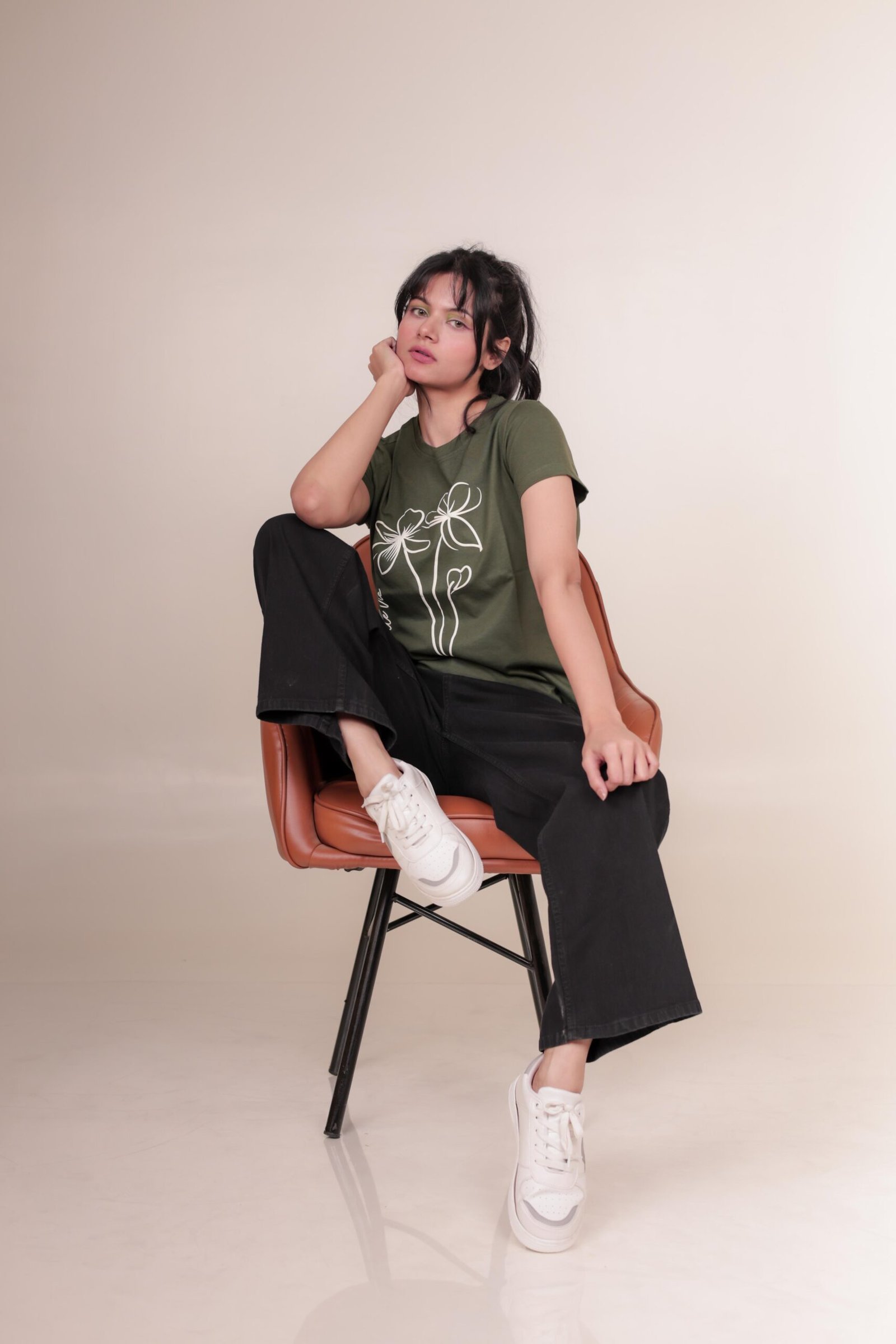 Casual studio portrait captured at Lens Grove Studio, a professional photo and video shoot rental space in Delhi, featuring a female model wearing a green graphic t-shirt, black pants, and white sneakers, posing against a minimal neutral background with soft, even lighting for lifestyle and portfolio photography.