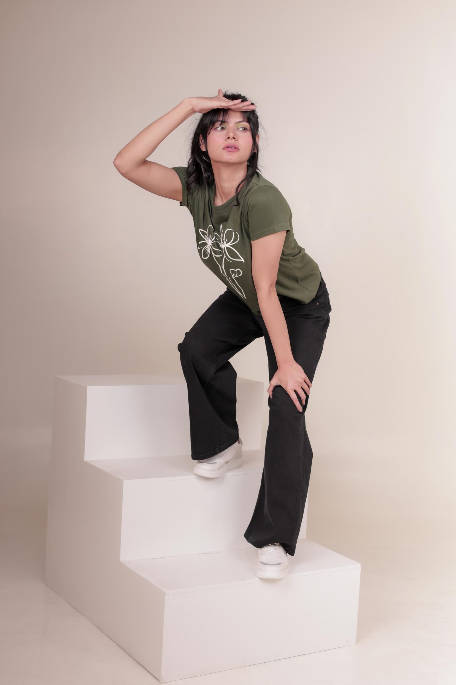 Casual studio portrait captured at Lens Grove Studio, a professional photo and video shoot rental space in Delhi, featuring a female model wearing a green graphic t-shirt, black pants, and white sneakers, posing against a minimal neutral background with soft, even lighting for lifestyle and portfolio photography.