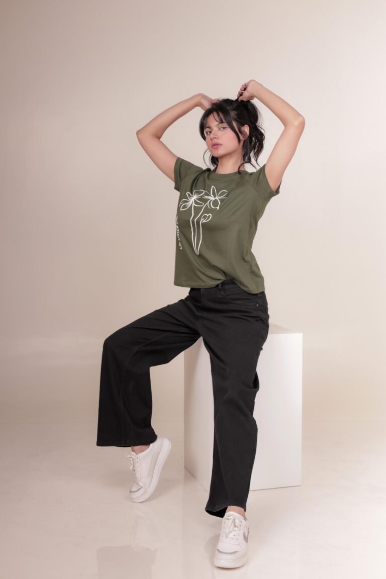 Casual studio portrait captured at Lens Grove Studio, a professional photo and video shoot rental space in Delhi, featuring a female model wearing a green graphic t-shirt, black pants, and white sneakers, posing against a minimal neutral background with soft, even lighting for lifestyle and portfolio photography.